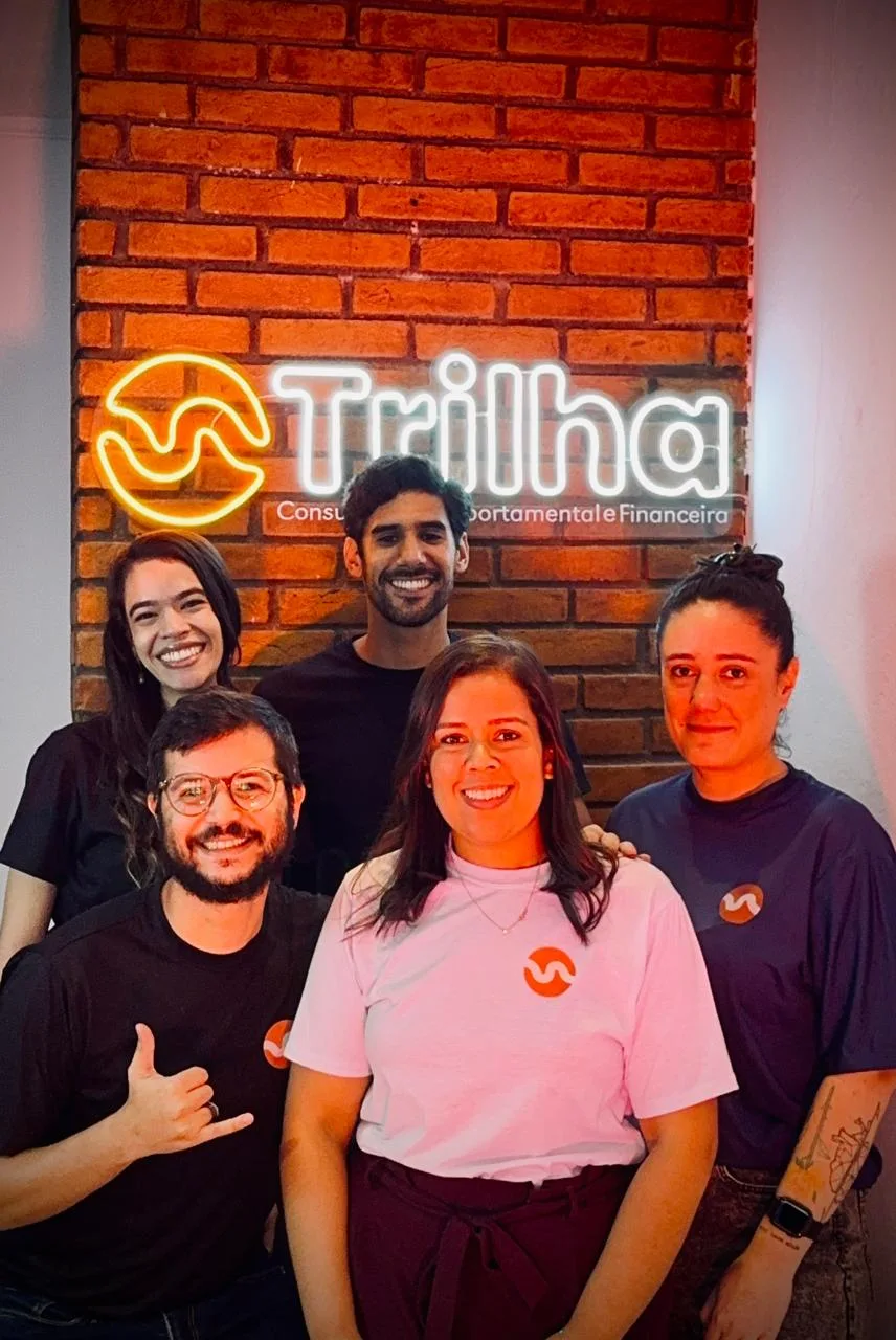 Integrantes da equipe da Trilha Consultoria em frente ao letreiro da marca no escritório